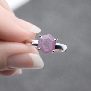 May include: A silver ring featuring a faceted, light purple gemstone. The gemstone is hexagonal in shape and set with four silver prongs. The ring is held against a gray background.