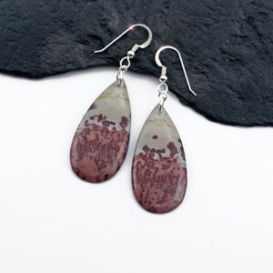 May include: A pair of teardrop-shaped earrings. The earrings feature a silver hook and a natural stone pendant. The stone has a mottled pattern of burgundy and gray-beige hues. The earrings are displayed on a white surface.