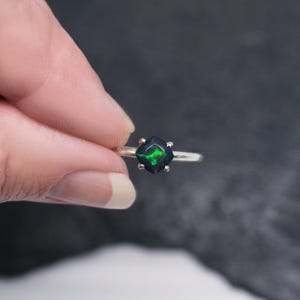 Peut inclure: Bague en argent ornée d'une opale noire taillée en carré, avec des reflets verts vifs. L'opale est sertie à quatre griffes. La bague est présentée sur un fond sombre. Dimensions de la pierre : environ 0,8 cm.