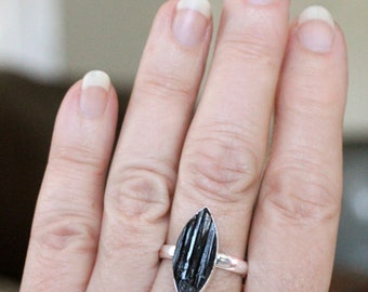 Puede incluir: Un anillo de plata con una piedra preciosa negra facetada de forma marquesa. El anillo está puesto en un dedo.