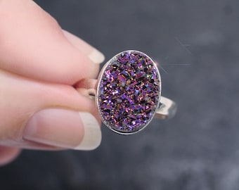 May include: Oval-shaped ring with a shimmering purple druzy stone set in a silver band. The druzy stone has a sparkling, crystalline texture, reflecting light with shades of purple and hints of other colors.