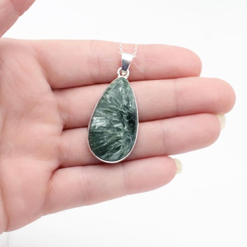 May include: A teardrop-shaped pendant with a silver-colored bezel and chain. The pendant features a green stone with a textured, fibrous pattern. The necklace is displayed against a white background.