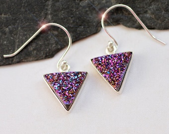 May include: A pair of silver-toned earrings with triangular, iridescent purple druzy stones. The earrings have a hook design for pierced ears. The druzy stones sparkle with shades of purple, blue, and pink, set in a silver frame.