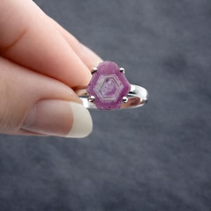 May include: A silver ring featuring a raw, hexagonal-shaped pink gemstone. The gemstone has a lighter, geometric pattern in the center. The ring is held against a dark gray background.