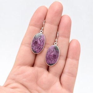 May include: A pair of oval-shaped drop earrings featuring polished purple and dark grey patterned stones set in silver-toned metal frames with simple hook-style ear wires for secure attachment to the earlobe.