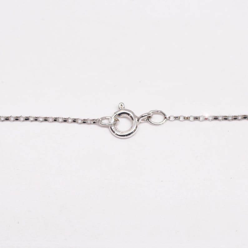 May include: Close-up of a silver necklace chain with a circular clasp. The chain is made of small, rectangular links. The clasp is a spring ring type, commonly used for jewelry. The background is plain white.