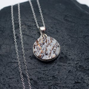 May include: A silver necklace featuring a round pendant with a speckled, light gray and brown stone. The pendant is set in a silver bezel and hangs from a delicate silver chain. The stone has a unique, natural pattern.