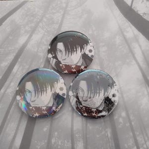 Puede incluir: Tres botones redondos con un personaje de estilo manga con cabello oscuro y una expresión seria. Cada botón tiene un esquema de color diferente, con variaciones sutiles en la cara y los detalles del personaje. Fondo borroso en escala de grises.