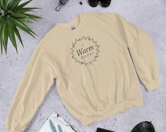Könnte beinhalten: Ein beigefarbenes Langarm-Sweatshirt mit Rundhalsausschnitt und einer dunkelgrünen, kreisförmigen Kranzgrafik in der Mitte der Brust. Im Inneren des Kranzes steht der Text "Warm & cosy" in einer dunkelgrünen Serifenschrift, ergänzt durch eine kleine Schleife.