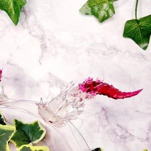 Könnte beinhalten: Eine durchsichtige Glaskuppel mit einer pinkfarbenen und weißen kristallartigen Struktur im Inneren. Die Kuppel steht auf einer weißen Marmorfläche mit grünen Efeu-Blättern drumherum.