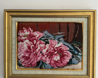 Op de afbeelding: Een ingelijst naaldkunstwerk met roze bloemen en groene bladeren. De bloemen staan in een vaas met een bruine achtergrond.