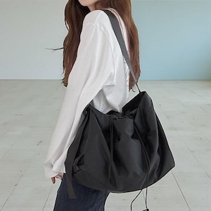 May include: A large black tote bag with a drawstring closure and long shoulder straps. The bag is made of a soft, flexible material and has a casual, relaxed style. The bag is designed to be worn over the shoulder.
