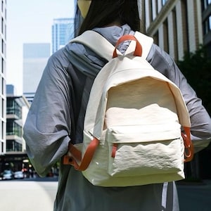 May include: A cream-coloured backpack with a large main compartment and a smaller front zipped pocket. The bag features brown leather-look straps and a top handle, with a textured fabric finish, worn on the back over a grey long-sleeved shirt.