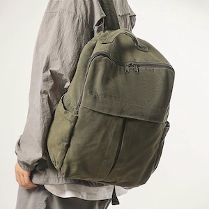 Pode incluir: Mochila de lona verde-oliva com vários bolsos e uma alça superior. A mochila tem formato retangular com um bolso com aba na frente e um compartimento principal com zíper. A bolsa é feita de tecido durável e texturizado.