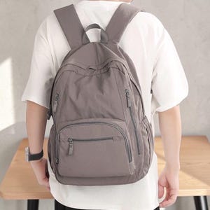 Könnte beinhalten: Ein einfarbiger taupefarbener Rucksack mit einem großen Hauptfach, einer kleineren Reißverschlusstasche auf der Vorderseite und zwei Seitentaschen. Der Rucksack verfügt über verstellbare Schultergurte, einen Tragegriff oben und passende Reißverschlüsse.