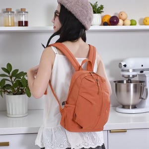 Puede incluir: Una mochila naranja con correas ajustables y un bolsillo frontal. La mochila tiene un compartimento principal con cremallera y un asa superior. La mochila está hecha de un material ligero y está diseñada para el uso diario.
