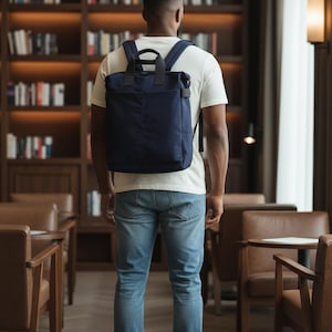 May include: A person wearing a white t-shirt and blue jeans, carrying a navy blue backpack with black handles. The person is standing in a library with bookshelves and leather chairs.