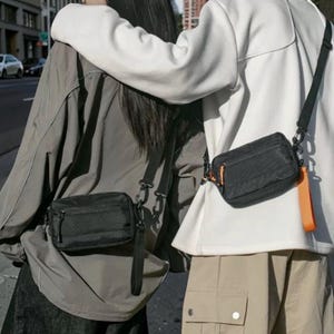 May include: Two black rectangular crossbody bags with zippered closures and orange pull tabs. Each bag features a main compartment and a smaller front pocket, attached to a black adjustable shoulder strap with metal hardware clips.