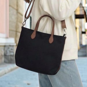 Può includere: Una borsa tote nera con manici in pelle marrone e una tracolla regolabile abbinata con clip in metallo color argento. La borsa ha una forma rettangolare strutturata con una base piatta, progettata per trasportare oggetti personali.
