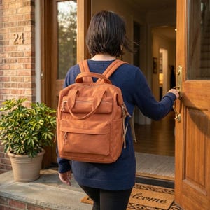 May include: A burnt orange canvas backpack featuring a large main compartment, a smaller front zippered pocket, two top carry handles, and adjustable shoulder straps. The bag has side pockets and matching fabric zipper pulls on all compartments.