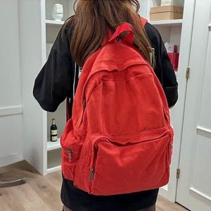 Może przedstawiać: Duży, jasnoczerwony plecak z wieloma kieszeniami i regulowanymi paskami. Plecak wykonany jest z wytrzymałej tkaniny i ma klasyczny design, odpowiedni do przenoszenia książek, laptopów lub niezbędnych rzeczy w podróży. Torba jest noszona na ramionach.