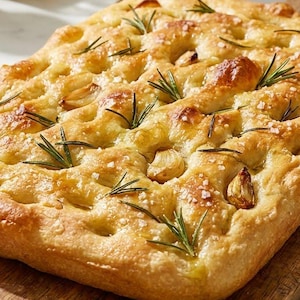 May include: A golden-brown focaccia bread topped with rosemary, garlic, and coarse salt, resting on a wooden cutting board. A small bowl of olive oil and a knife are nearby. The bread has a dimpled texture.