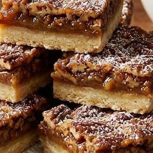 Puede incluir: Una pila de barras de pastel de nuez pecana espolvoreadas con azúcar glas. Las barras tienen una base dorada y mantecosa y un rico relleno de nuez pecana. El relleno es de color marrón oscuro, con nueces pecanas visibles. Las barras están cortadas en cuadrados.