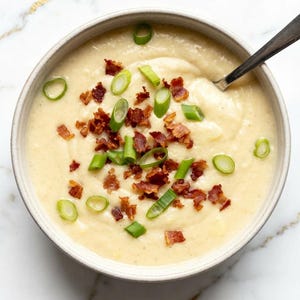 May include: A round, light-colored ceramic bowl filled with a creamy, off-white potato soup. The soup is topped with scattered pieces of crispy, dark brown bacon bits and bright green sliced scallions. A silver metal spoon rests in the bowl.