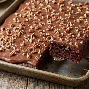 Könnte beinhalten: Ein rechteckiger Schokoladenkuchen in einer Metallbackform. Der Kuchen ist mit Schokoladenfrosting bedeckt und mit gehackten Pekannüssen belegt. Eine Scheibe wird mit einem Metallspatel angehoben. Der Kuchen steht auf einer Holzoberfläche.
