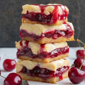 May include: A stack of four cherry pie bars with a golden crust and a vibrant red cherry filling. Fresh cherries are scattered around the base of the stack. The bars have a white, creamy topping and a drizzle of red cherry sauce.