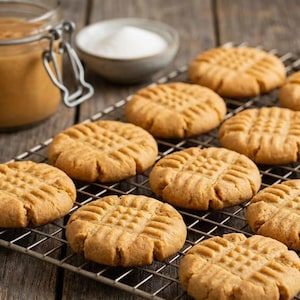 Puede incluir: Primer plano de galletas de mantequilla de cacahuete sobre una rejilla metálica. Las galletas son redondas, de color marrón dorado y tienen un patrón entrecruzado en la parte superior. Un tarro de mantequilla de cacahuete y un cuenco pequeño de azúcar están en el fondo.