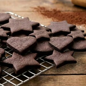 Può includere: Primo piano di biscotti al cioccolato a forma di stella, cuore e cerchio su una griglia di raffreddamento in metallo. I biscotti sono di un ricco colore marrone e si trovano su una superficie di legno. La polvere di cacao è cosparsa sullo sfondo.