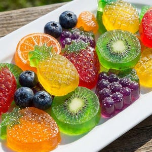 Könnte beinhalten: Ein weißer Teller mit bunten, fruchtförmigen Gummibonbons. Das Sortiment umfasst Erdbeer-, Orangen-, Kiwi-, Ananas- und Traubengummis sowie frische Blaubeeren. Die Süßigkeiten sind auf einer Holzoberfläche angeordnet.