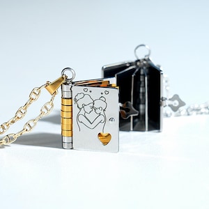 Puede incluir: Collar con colgante en forma de libro pequeño, plateado y dorado. El libro tiene una cadena y un cierre dorados. La portada presenta una imagen grabada de dos personas abrazándose, con un corazón recortado.