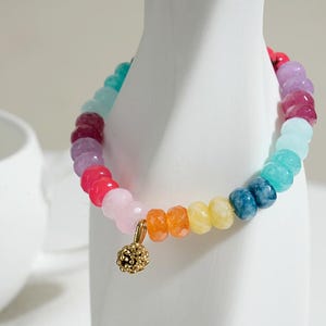 Puede incluir: Una pulsera de cuentas coloridas con un dije dorado. La pulsera presenta un arcoíris de colores, incluyendo rosa, rojo, morado, azul, verde, amarillo y naranja. El dije es una pequeña bola dorada.
