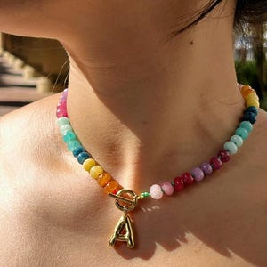 Peut inclure: Un collier de perles colorées avec un pendentif initial "A" de couleur or. Le collier présente un arc-en-ciel de perles, comprenant des nuances de rouge, rose, jaune, vert et bleu. Le fermoir est de couleur or.
