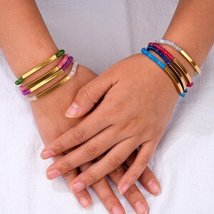 Puede incluir: Brazaletes de metal dorado con detalles de cuentas de colores. Los brazaletes presentan un diseño elegante y minimalista con un acabado pulido. Las cuentas son en tonos rosa, verde, blanco, azul y rojo. Los brazaletes están apilados en la muñeca.