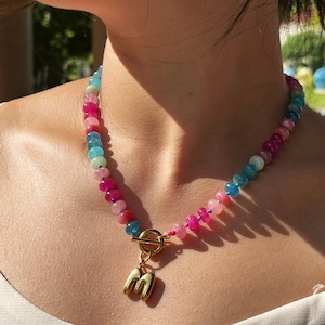Könnte beinhalten: Eine farbenfrohe Perlenkette mit einem goldfarbenen Verschluss und einem goldenen Anhänger mit dem Buchstaben "M". Die Perlen sind in Rosa-, Blau- und Grüntönen gehalten. Die Halskette wird um den Hals getragen.
