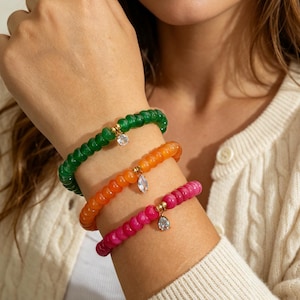 May include: Three stretch bracelets featuring round, translucent beads in vibrant green, orange, and hot pink. Each bracelet includes a single gold-toned metal spacer bead holding a clear, faceted, teardrop-shaped crystal charm dangling from the center.