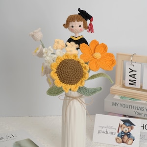 Könnte beinhalten: Ein handgefertigter Häkelstrauß in einer weißen Vase mit einer Sonnenblume, einer orangefarbenen Blume und anderen weißen und gelben Blumen. Oben befindet sich eine Häkelpuppe mit Abschlussmütze. Eine Karte lautet "Herzlichen Glückwunsch zum Abschluss" mit einem Teddybär mit Abschlusskappe.