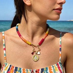 May include: A necklace featuring a strand of multi-colored beads in a rainbow gradient. The necklace includes a gold-toned star-shaped clasp with three hanging charms: a small dark red teardrop, a small white sun, and a round gold coin with the letter A.