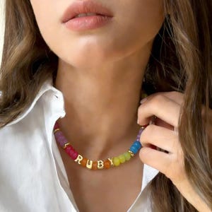 Könnte beinhalten: Eine farbenfrohe Perlenkette mit dem Namen "RUBY" in goldenen Buchstaben. Die Perlen sind in einem Regenbogenmuster angeordnet, mit Farben wie Rot, Rosa, Orange, Gelb, Grün und Blau. Die Kette wird über einem weißen Hemd getragen.