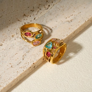 Könnte beinhalten: Zwei goldfarbene Ringe mit mehrfarbigen Edelsteinen. Ein Ring hat ein mehrschichtiges Banddesign mit ovalen rosa, blauen und hellrosa Steinen. Der zweite Ring ist ein breiteres Band, besetzt mit verschieden geformten blauen, rosa, orangen und grünen Steinen.