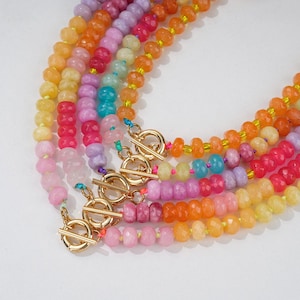 Puede incluir: Una colección de collares de cuentas coloridas en tonos rosa, amarillo, naranja y morado. Cada collar presenta un cierre de palanca dorado. Las cuentas son facetadas y están ensartadas en un cordón colorido.
