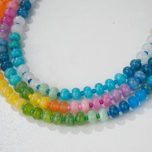 Puede incluir: Primer plano de tres hilos de collares de cuentas de colores. Las cuentas son en tonos de amarillo, verde, naranja, rosa, azul y morado. Los collares están sobre una superficie blanca.