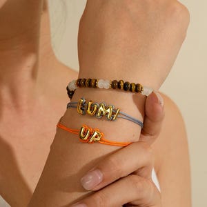 Könnte beinhalten: Ein Set aus drei zusammen getragenen Armbändern. Das obere Armband besteht aus abwechselnden braunen Tigerauge- und weißen Perlen. Das mittlere graue Kordelarmband zeigt goldfarbene Buchstaben, die LUMI buchstabieren. Das untere orangefarbene Kordelarmband trägt die goldfarbenen Buchstaben UP.