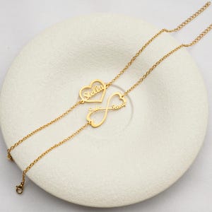 Könnte beinhalten: Zwei goldfarbene Halsketten mit personalisierten Namensanhängern. Eine Kette hat einen herzförmigen Anhänger mit dem Namen "Stella", die andere ein Unendlichkeitszeichen mit "Edie & Penny". Die Halsketten liegen auf einer weißen Oberfläche.