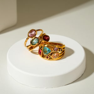 May include: Two gold-toned rings featuring an array of multi-colored gemstones. The larger ring has a wide band with various shapes including a central triangular blue stone, while the smaller ring features oval pink and blue stones.