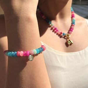 Könnte beinhalten: Ein farbenfrohes Perlenarmband- und Halsketten-Set. Das Armband hat einen Anhänger mit dem Buchstaben "A". Die Halskette hat einen goldfarbenen Verschluss und einen Anhänger. Die Perlen sind in Rosa-, Blau- und Grüntönen gehalten.