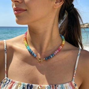 Puede incluir: Un collar de cuentas de colores con un cierre dorado. El collar presenta un degradado de colores del arcoíris, incluyendo rojo, naranja, amarillo, verde, azul y morado. El collar lo lleva una persona en un día soleado cerca del mar.